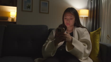 Woman-Spending-Evening-At-Home-Sitting-On-Sofa-With-Mobile-Phone-Scrolling-Through-Internet-Or-Social-Media-4